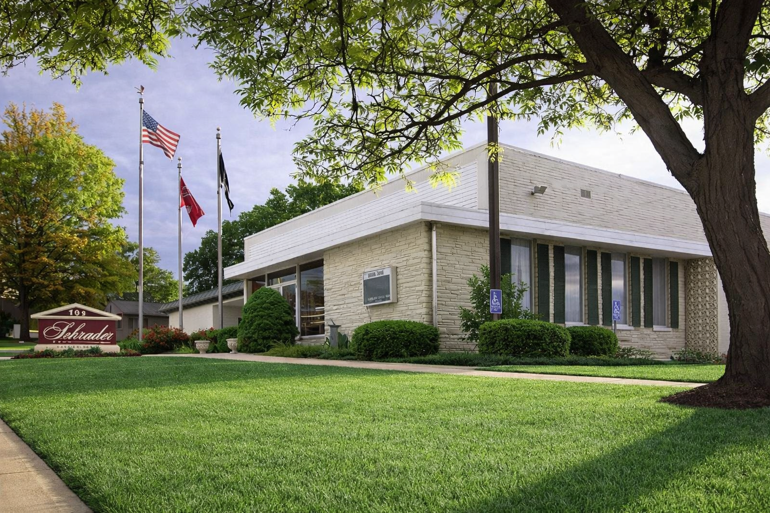 Brick office building with flags, a sign, and a manicured lawn under trees