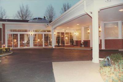 Covered entrance to a building at dusk with lit windows and a driveway under a white canopy