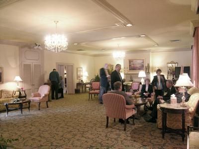 Elegant lounge with chandeliers, patterned carpet, and several people seated and standing near tables with lamps.