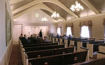 Interior of a church or chapel with pews, chandeliers, and a few people seated near the front.