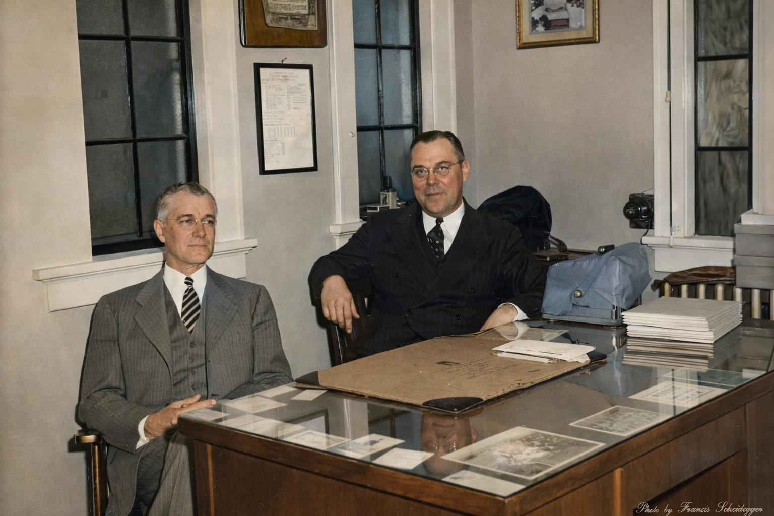 Two men seated at a desk in an office, with papers and framed items on the walls.