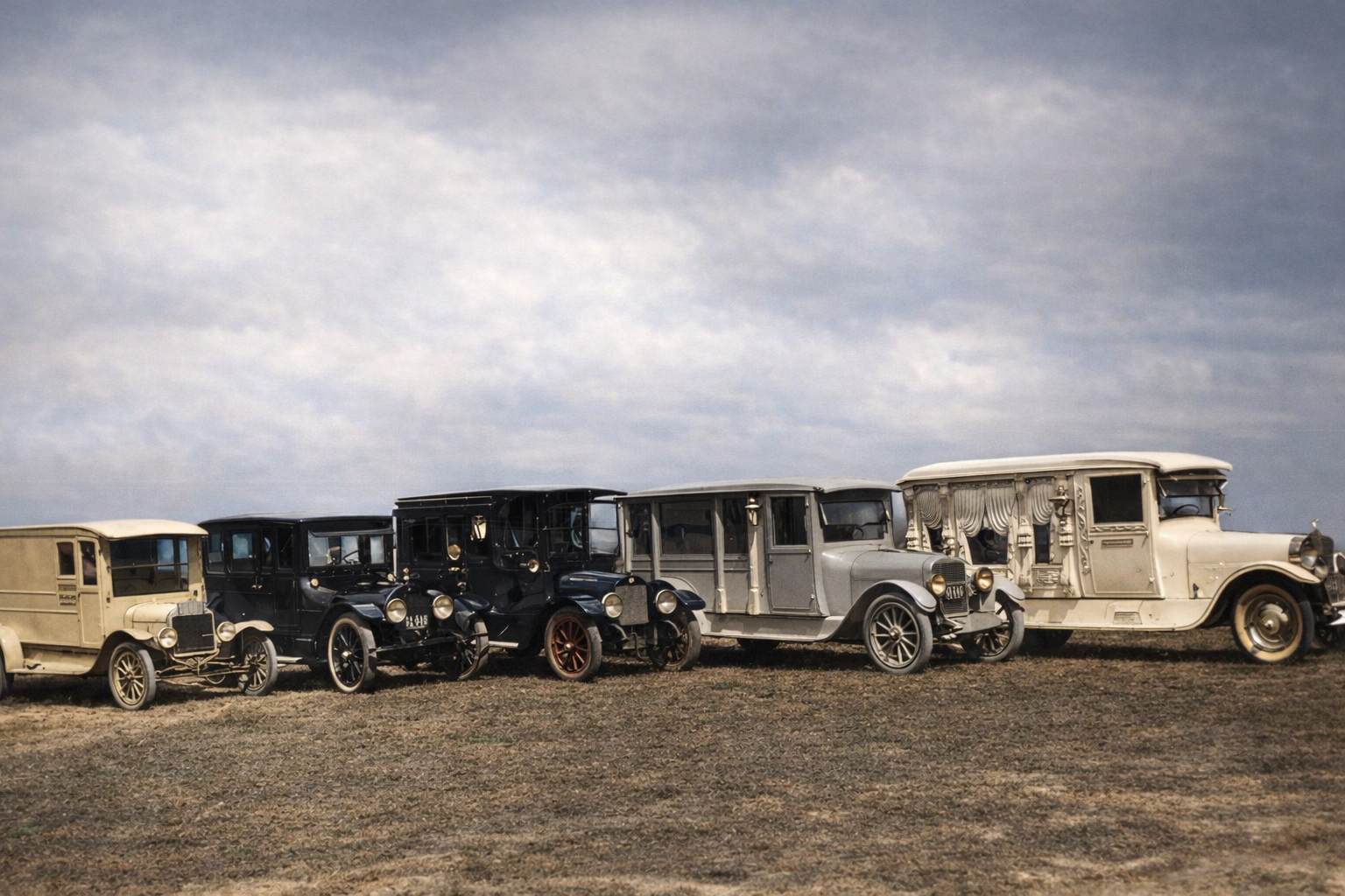 A line of vintage cars parked on a dry field under a cloudy sky