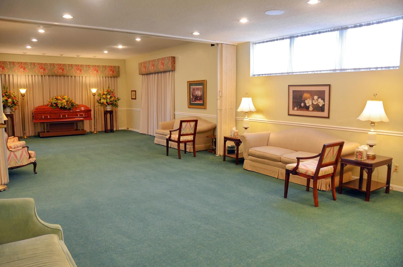 Quiet waiting room with green carpet, beige sofas, chairs, lamps, and floral curtains