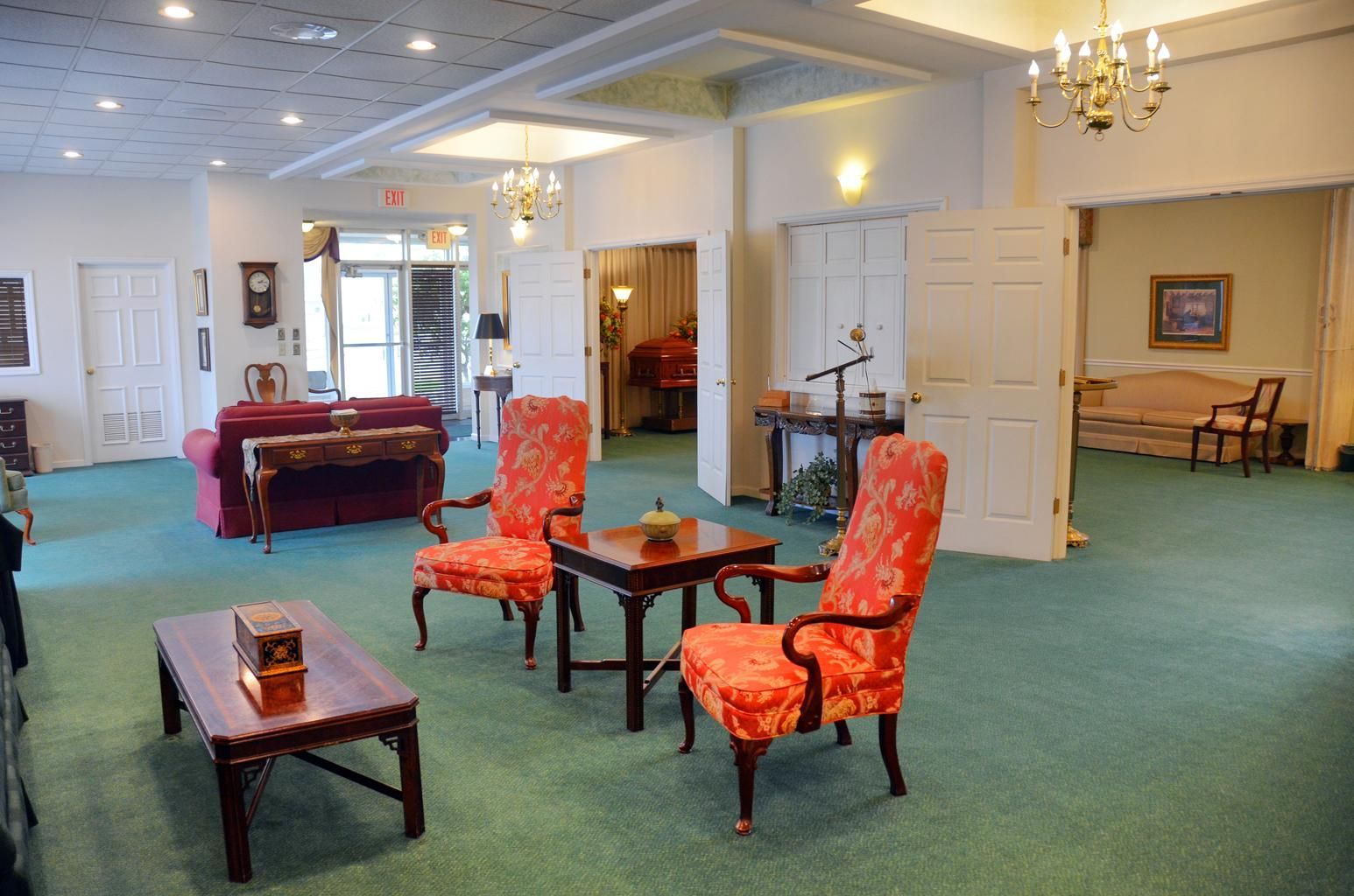 Spacious lounge with green carpet, red chairs, tables, chandelier lights, and a sofa.