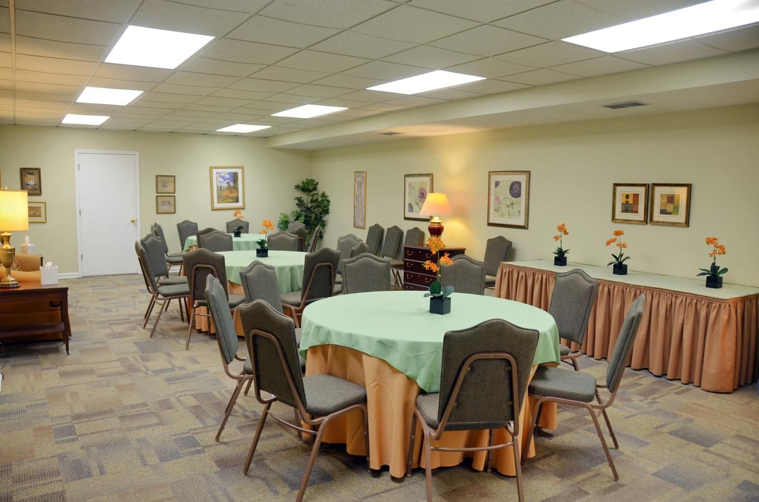 Community dining room with round tables, chairs, and serving tables under bright fluorescent lights.