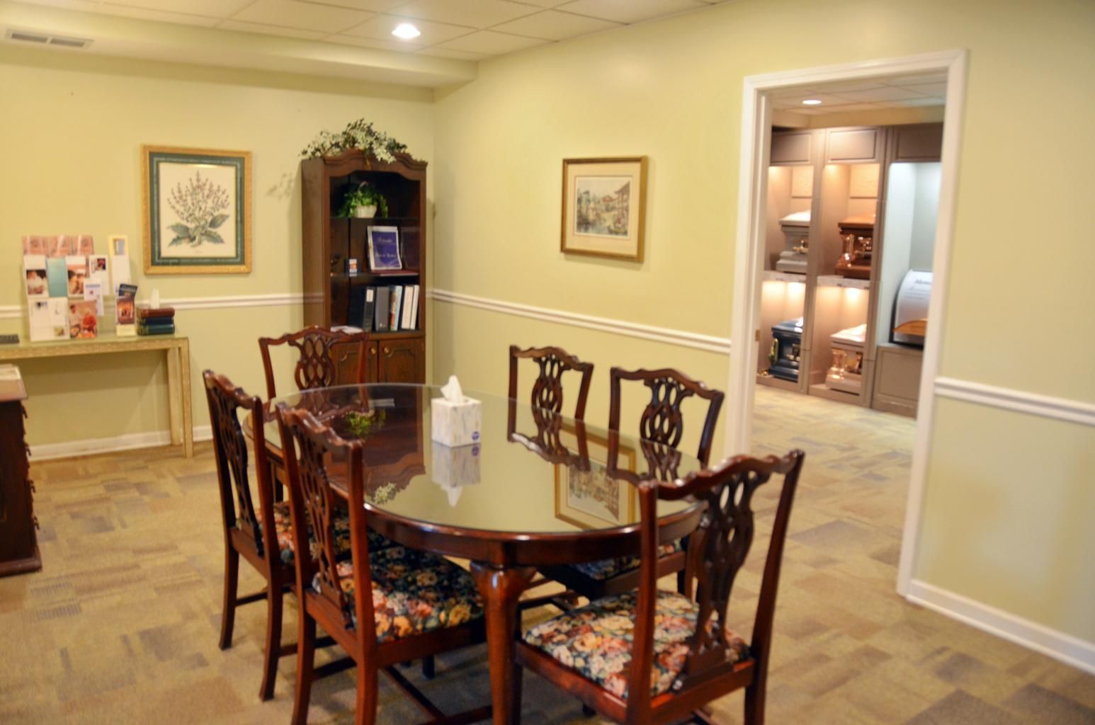 Dining room with glass table and carved chairs, warm beige walls, and a doorway to a lit room beyond.