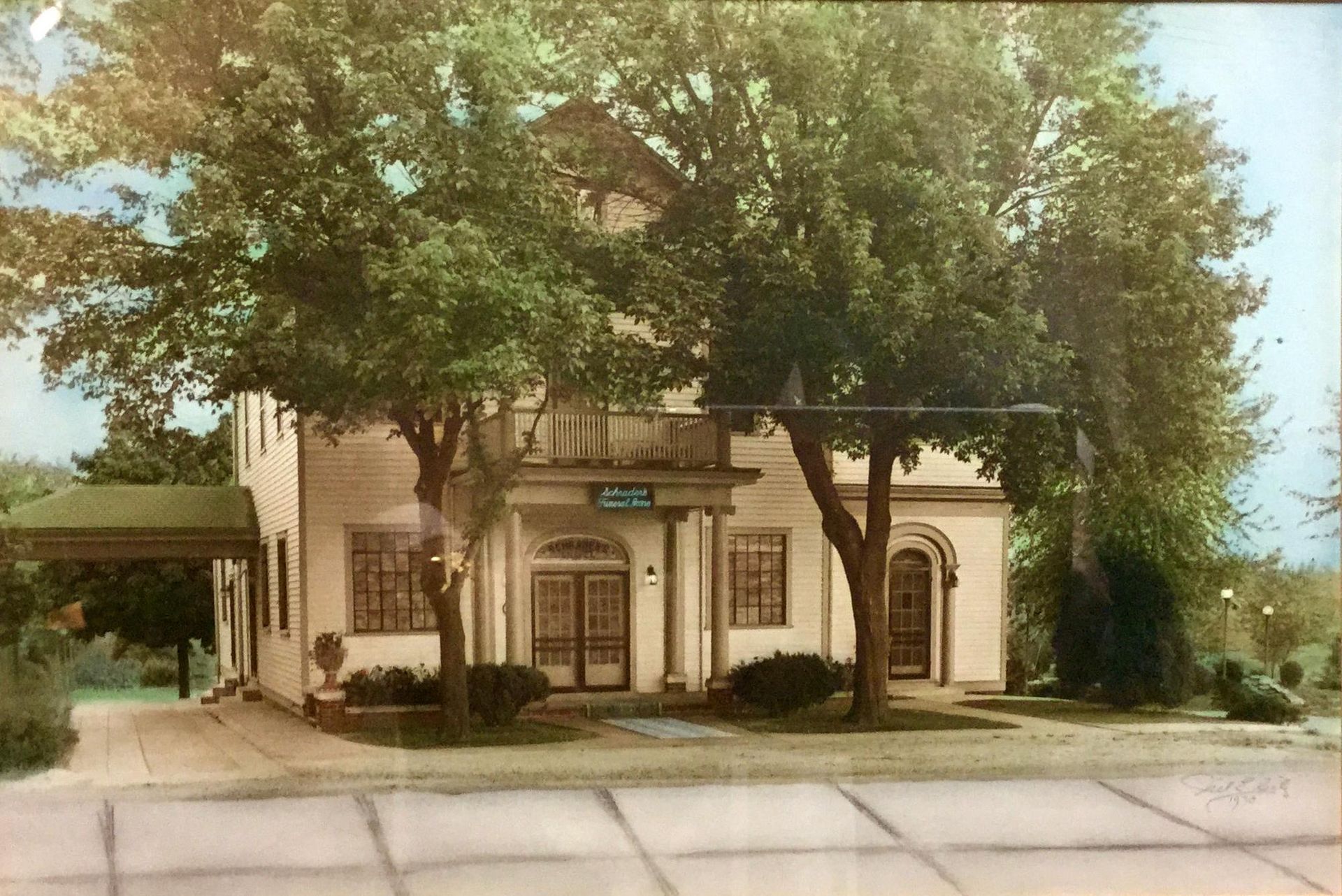 Tree-shaded beige building with arched windows and storefronts along a quiet street
