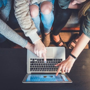 Three people point at a laptop screen while sitting around a black table.
