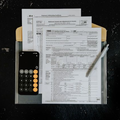 A calculator and pen sit on top of several IRS tax forms contained in a tan folder against a dark, textured background.