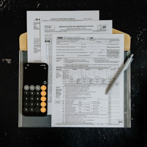 A calculator and pen sit on top of several IRS tax forms contained in a tan folder against a dark, textured background.