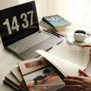 Hands hold an open book on a desk with a stack of books, a laptop displaying