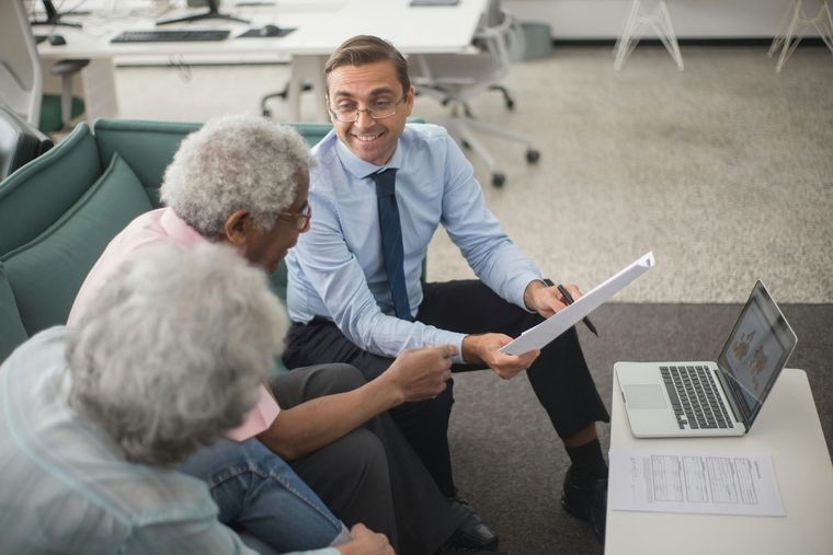 A professional advisor in a dress shirt and tie reviews documents with two clients seated together on a sofa.