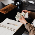 A person wearing a brown sweater counts a stack of US dollar bills at a desk next to an open planner and a pen.