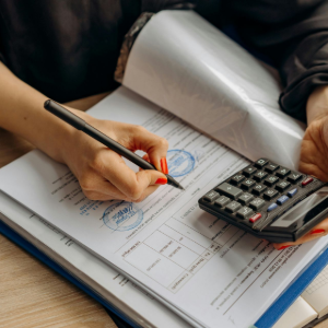 A person writes on a document with a pen while holding a calculator over a desk with folders.