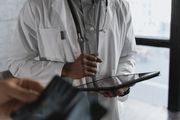 A person in a white lab coat with a stethoscope around their neck holds a digital tablet.