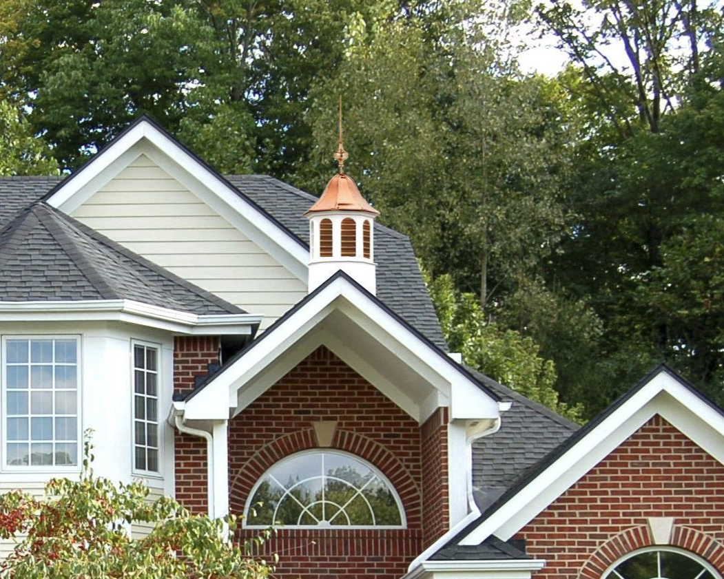 Brick house with black roof, white trim, and copper-topped cupola against a backdrop of green trees.