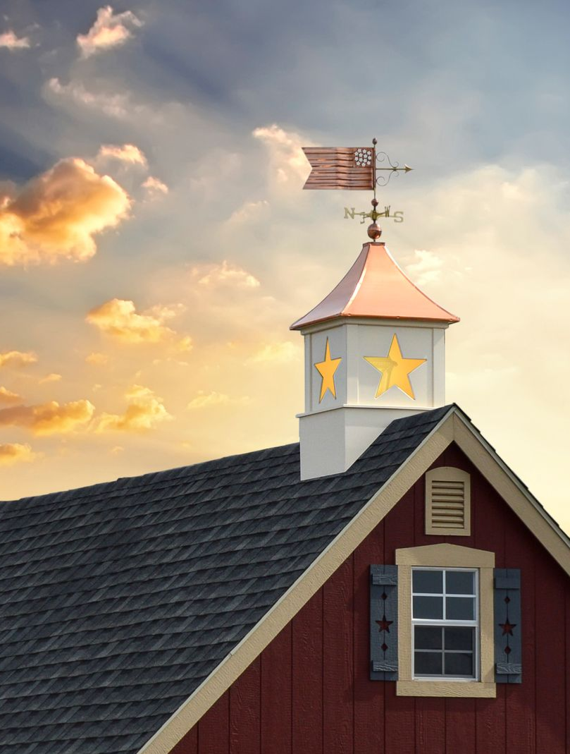 Red barn rooftop with copper-topped cupola, weather vane, and window, set against a sunset sky.