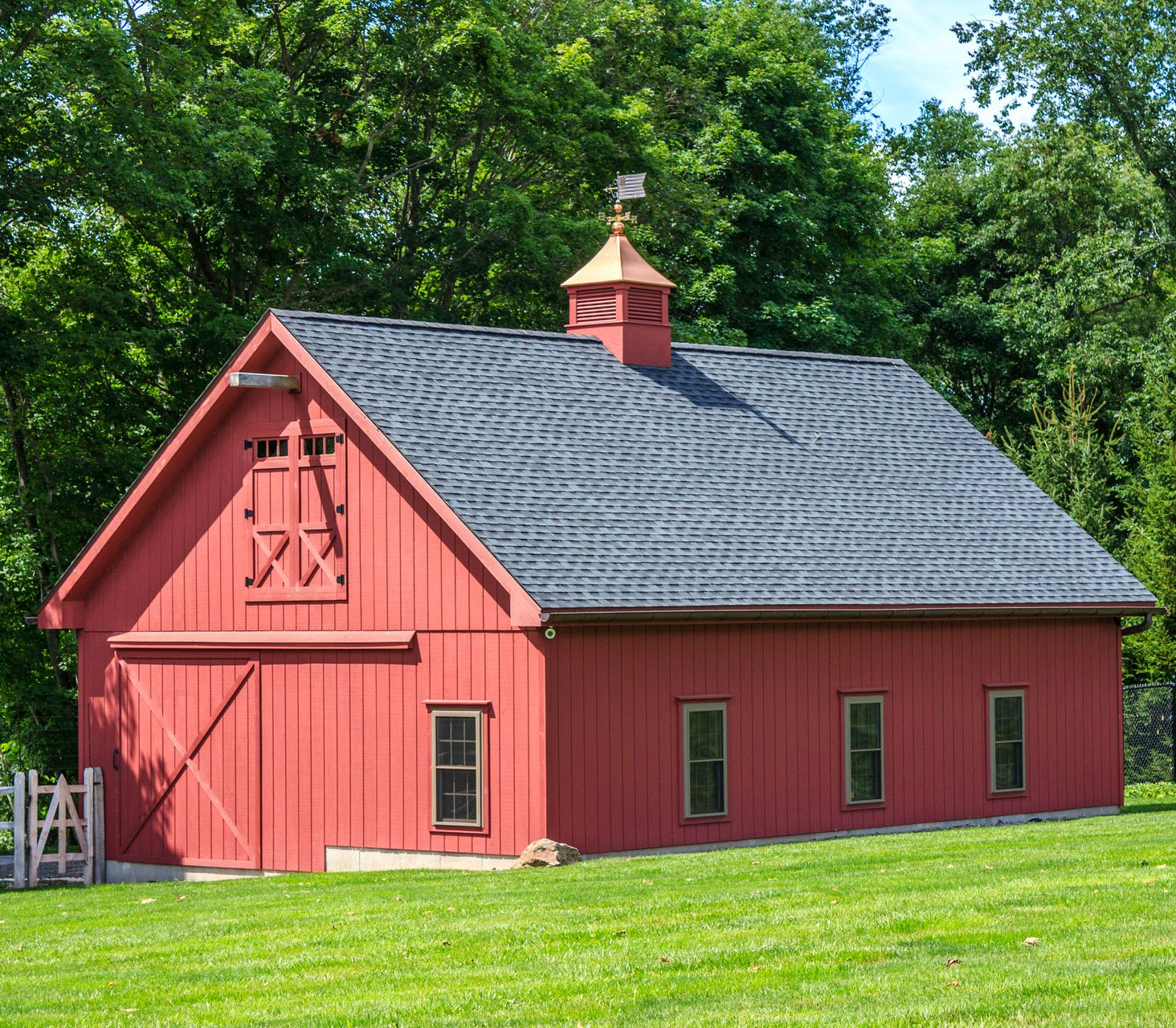 Red barn with a dark gray roof and weather vane, set in a grassy area with trees.
