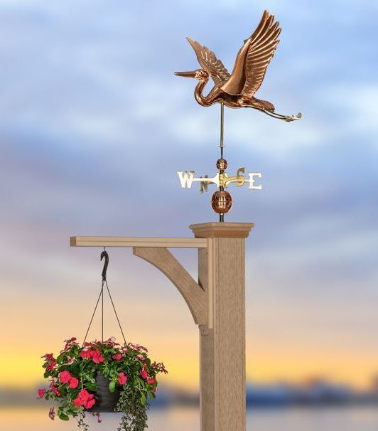 Copper heron weathervane atop a wooden post with hanging flower basket; dusk sky background.