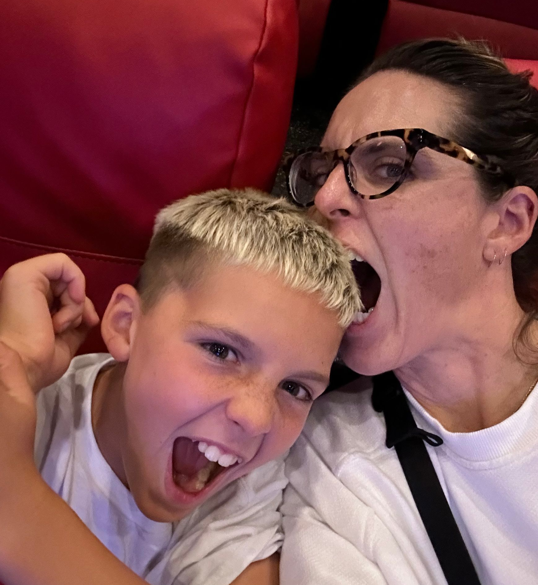 Two people making playful faces against red seats, one wearing glasses and a white shirt