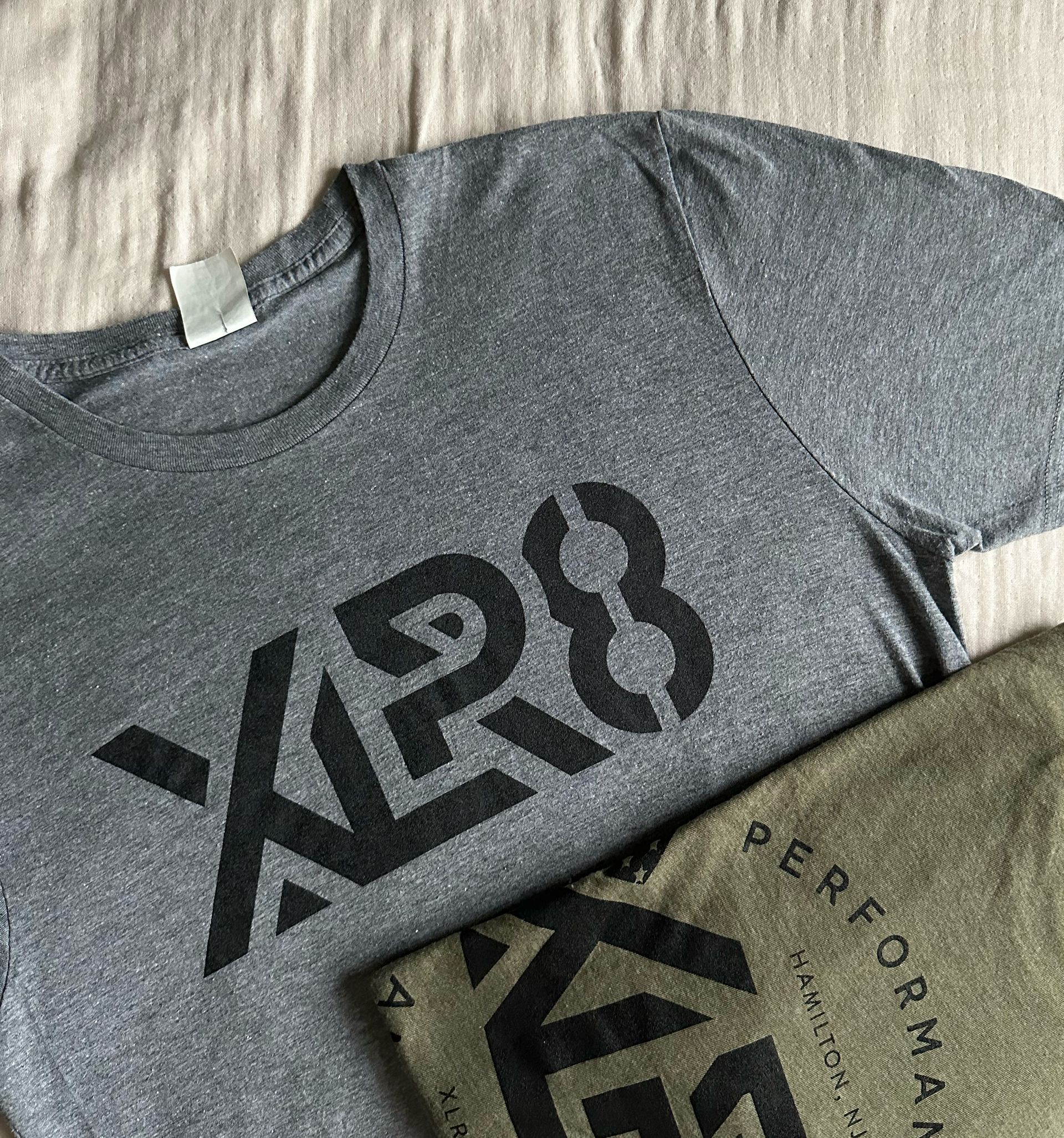 Two folded T-shirts on fabric, one gray and one olive green with bold black graphics.