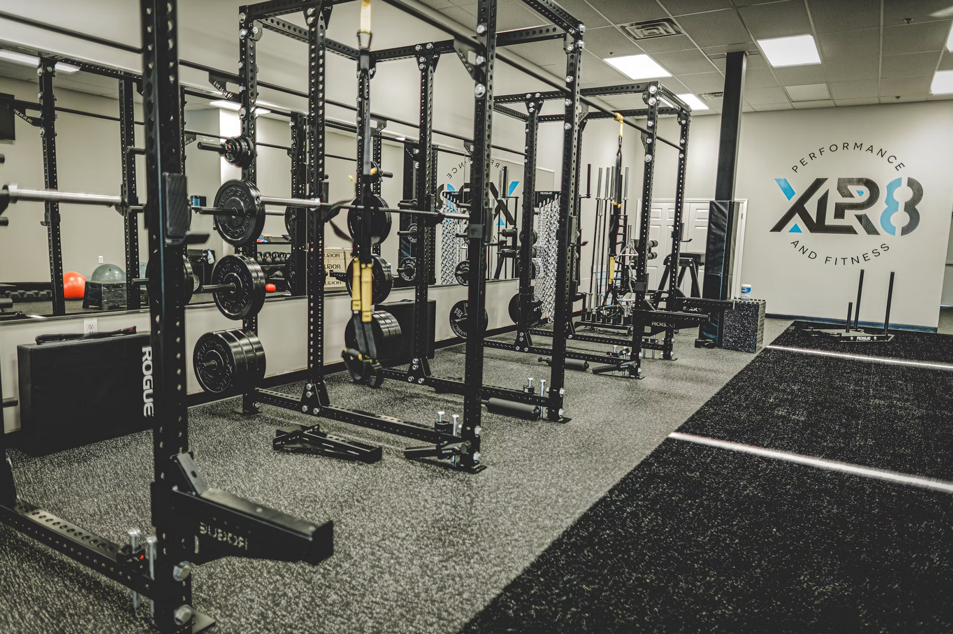 Indoor gym with black squat racks, weights, and turf flooring under bright lights
