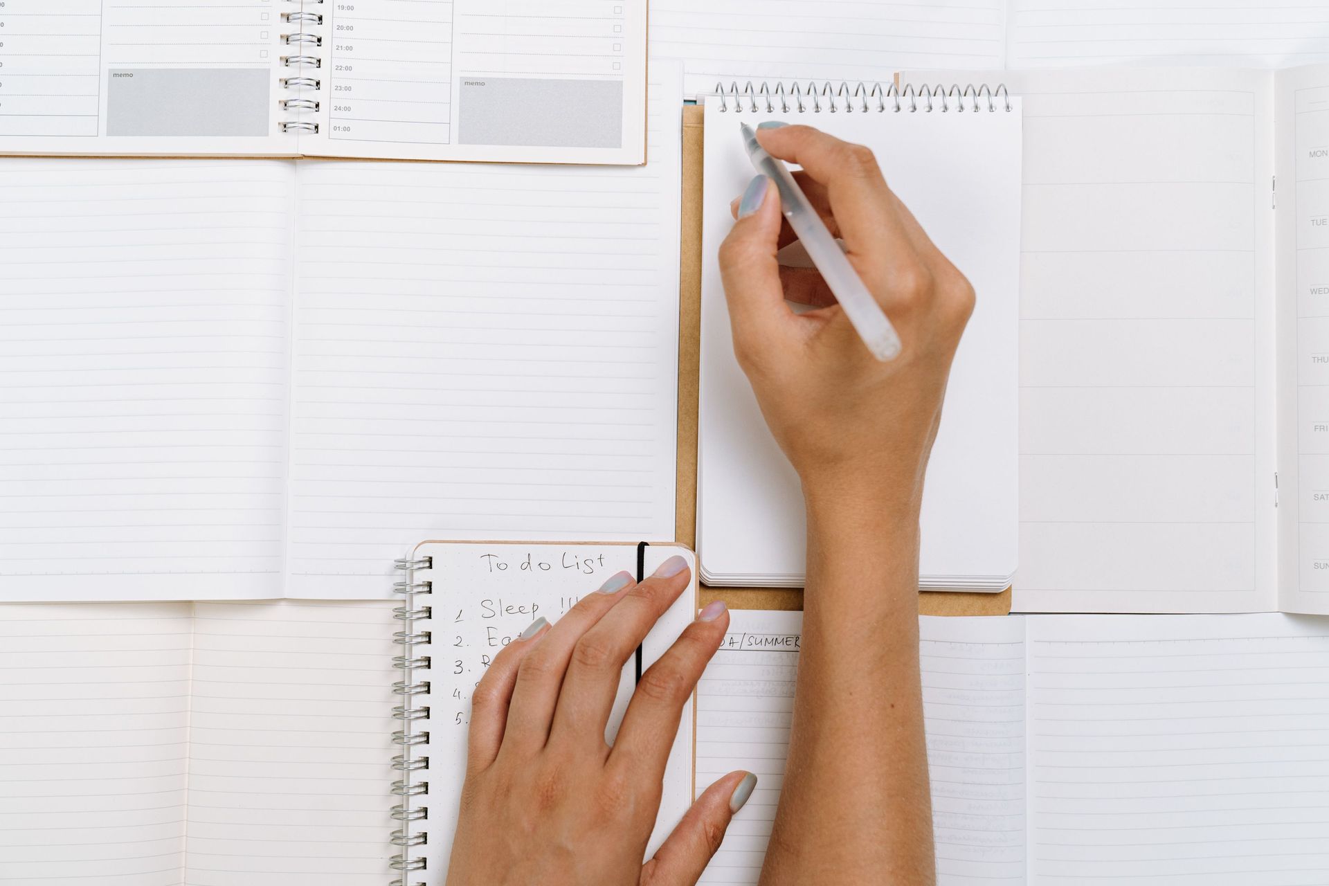 Hands writing notes in a small spiral notebook on a white desk with open planners and papers.