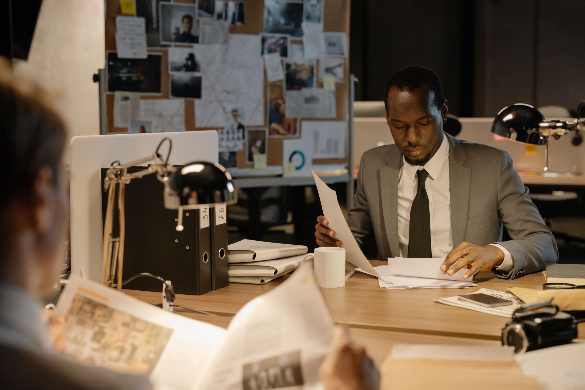 Man in a gray suit reviews papers at a cluttered office desk with lamps and a bulletin board behind him