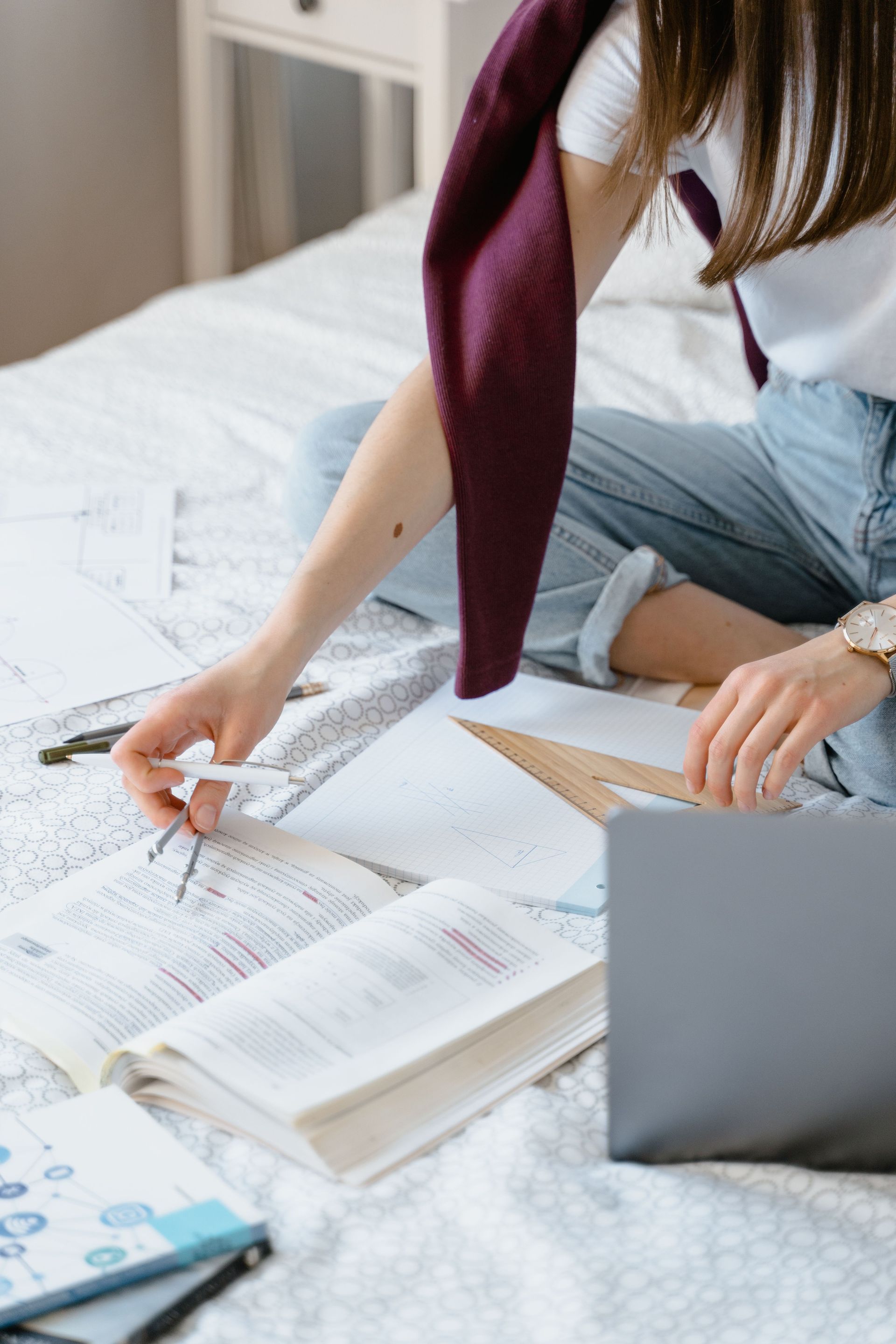 Person studying with open books and laptop on a bed, writing on papers with a pen.