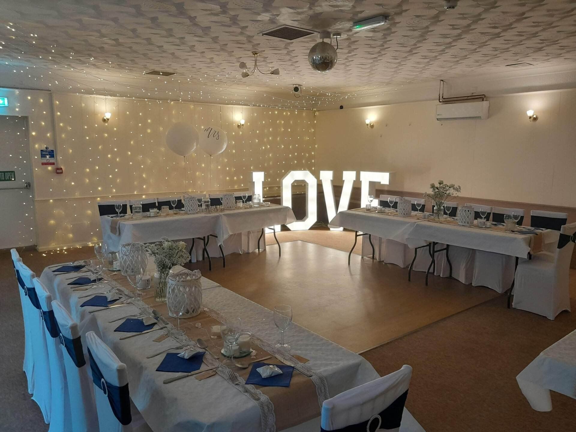 Four foot LOVE letter lights. Set up in a hall for a wedding, with sparkling lights and tables set up for a wedding meal.