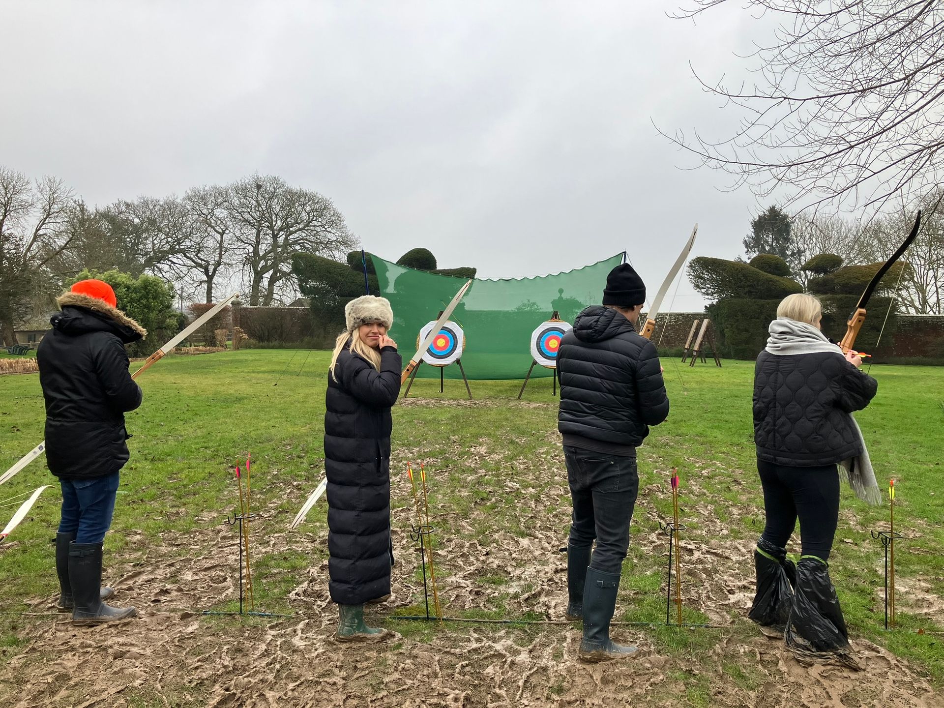 Archery bows and arrows set up in a line on a field with 2  targets in the distance with 4 people holding bows