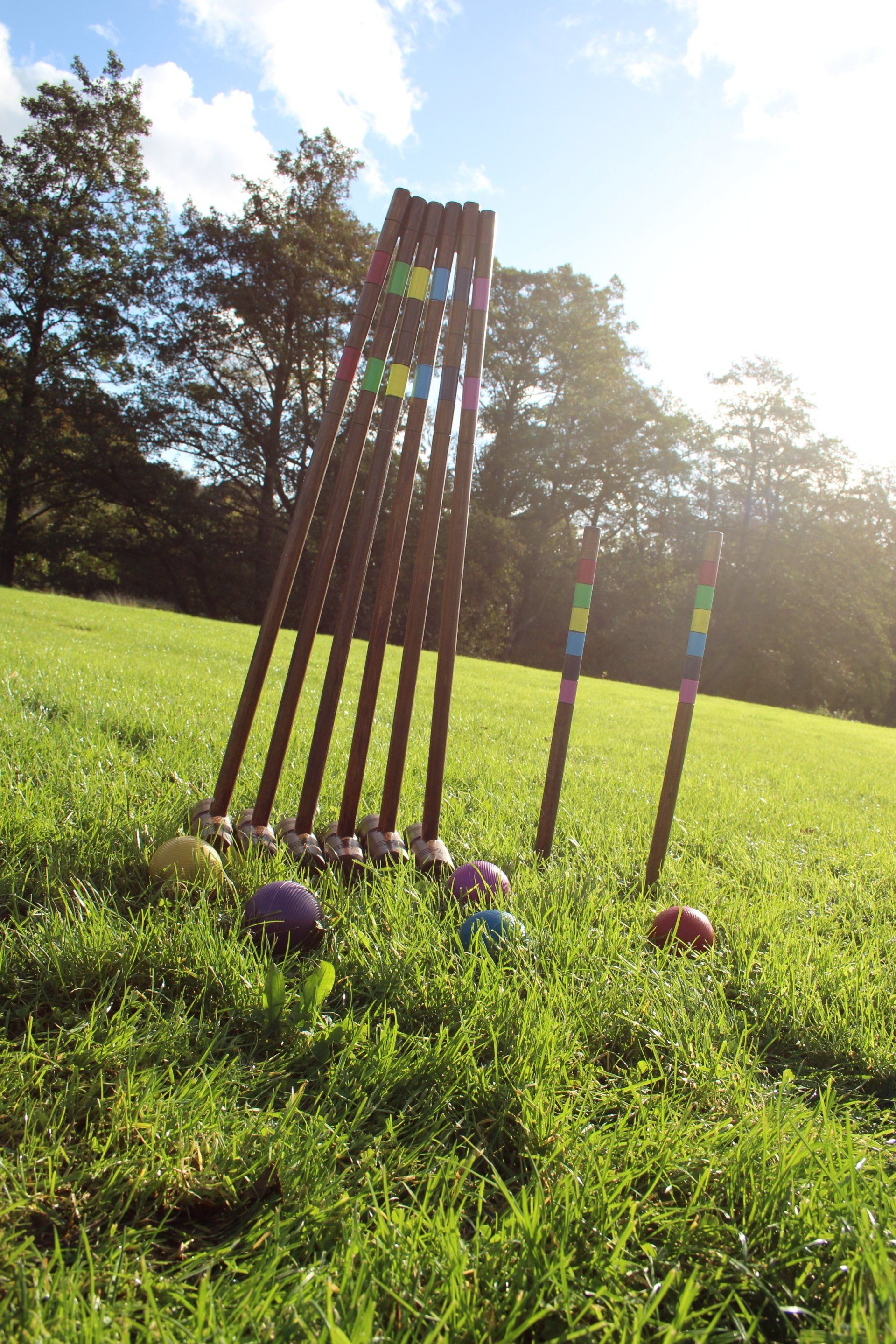 6 croquet mallets stood up vertically next to each other on grass, with two croquet sticks and 6 balls on the grass in front.
