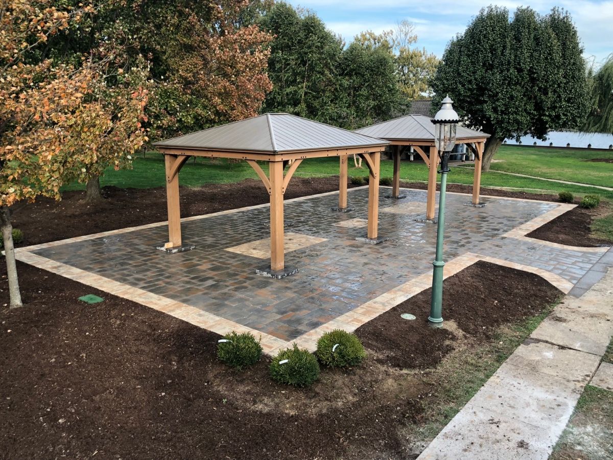 Stone-paved park pavilion with three wooden shelters, lamps, and surrounding pathways and shrubs