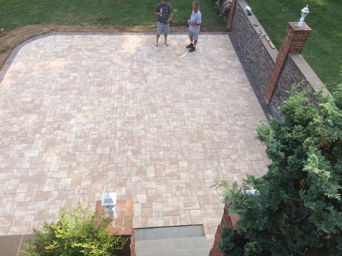 Two people standing on a large beige paved patio beside a brick wall and greenery.