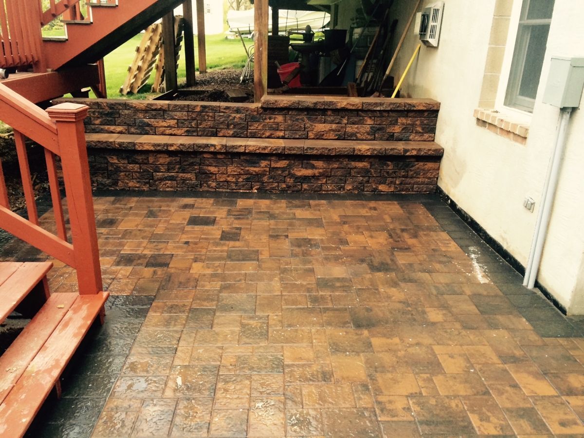 Brick patio leading to wooden steps and a porch, with a house wall on the right.