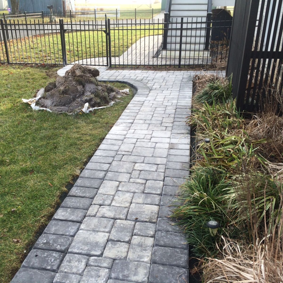 Brick walkway leading to a gated entrance beside a lawn and landscaped garden.