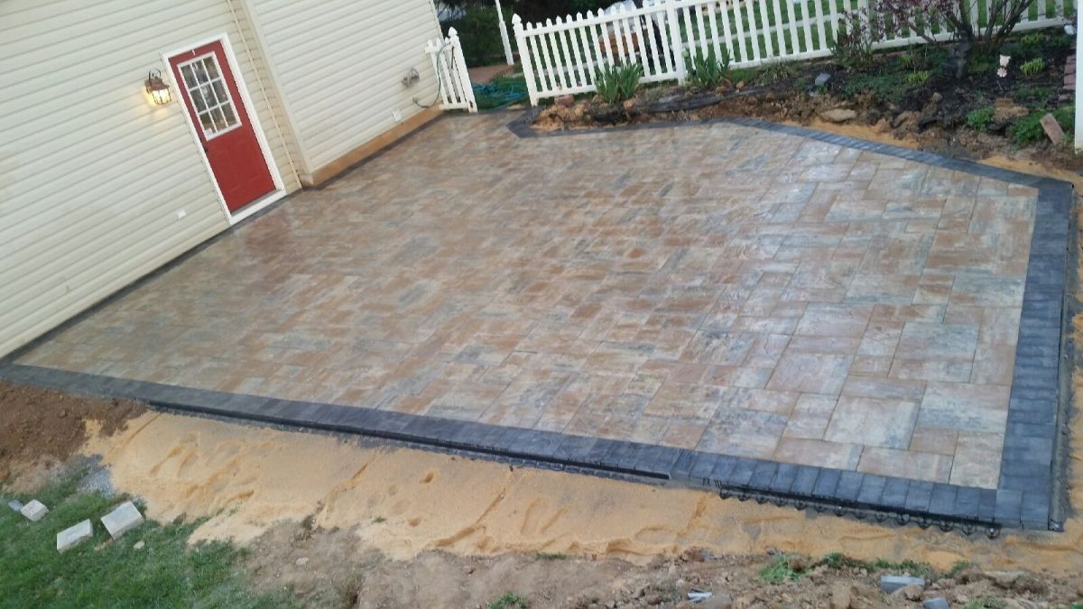 Empty brick patio beside a house, bordered with dark pavers and a small grassy edge