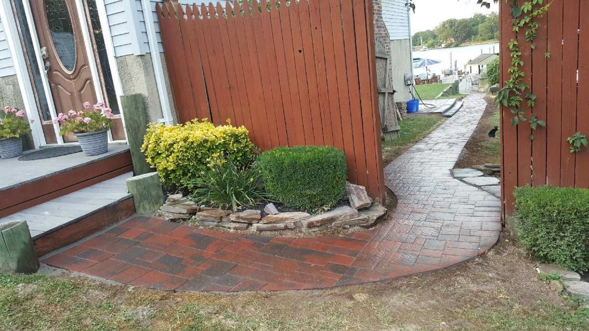 Brick walkway beside a red fence and garden beds leading to a waterfront path