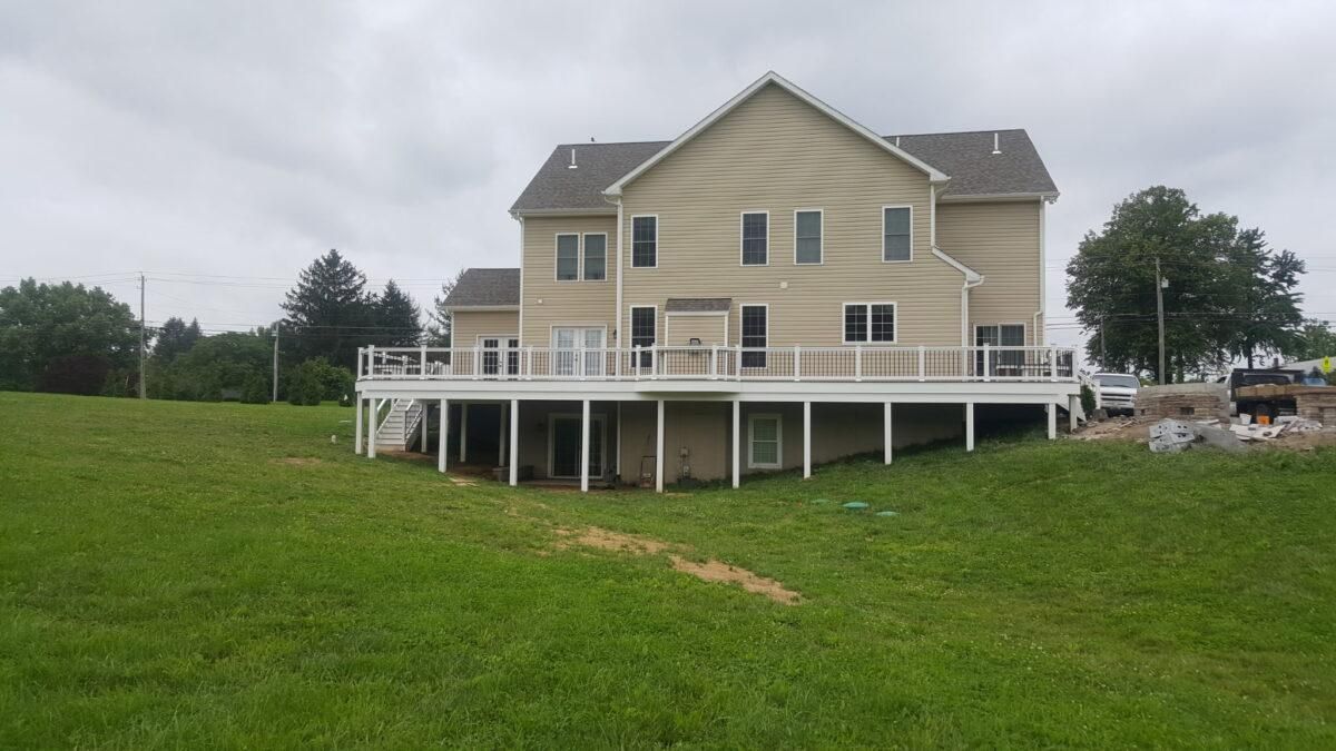 Large beige house with a wraparound deck on a grassy hill under cloudy skies
