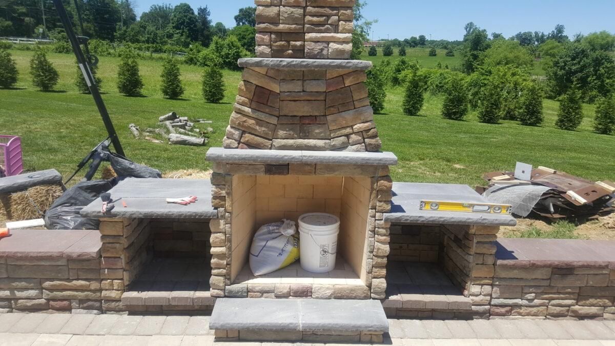 Stone outdoor fireplace with central chimney and built-in side shelves in a landscaped yard