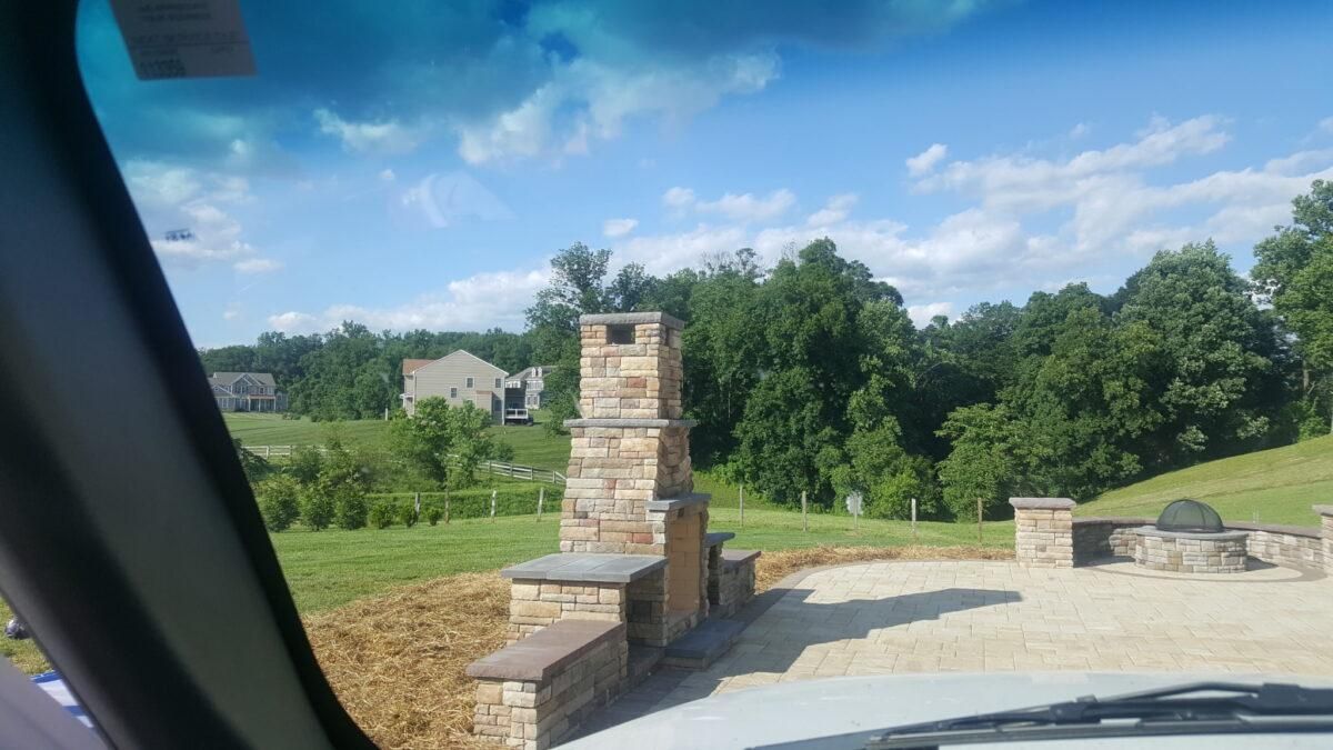 Stone fireplace and low wall beside a paved driveway overlooking green trees under a blue sky.