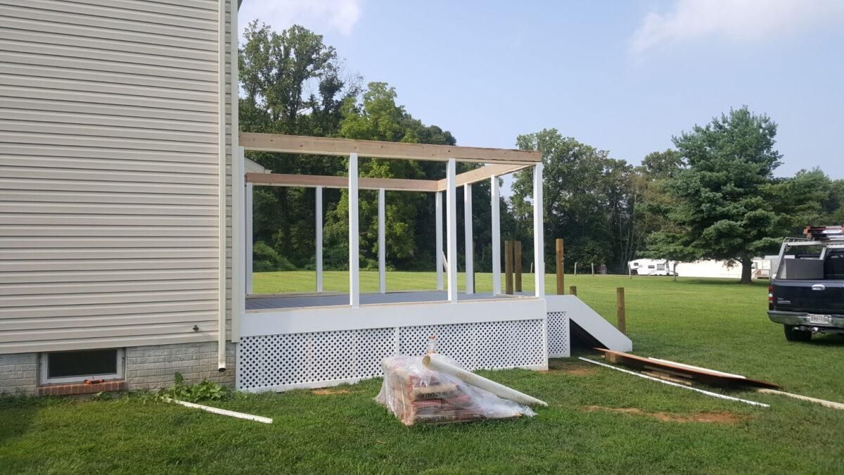 Porch under construction beside a beige house, with white posts, a tarp on the lawn, and a truck nearby.