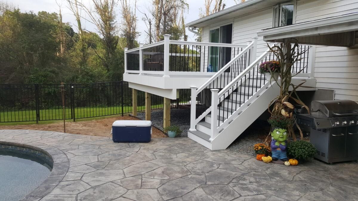 Backyard patio with stone paving, raised deck stairs, and a pool at left beside a white house.