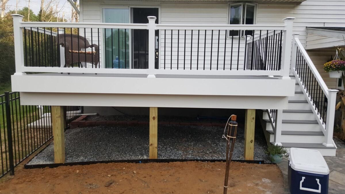 Elevated white deck with railing and stairs beside a house, with gravel and a dirt yard below.