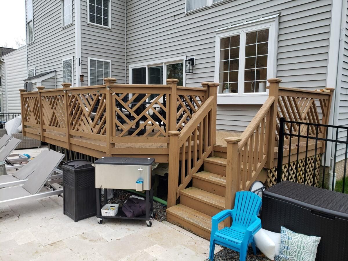 Wooden backyard deck with stairs beside a gray house, black railing, and outdoor chairs in a snowy area
