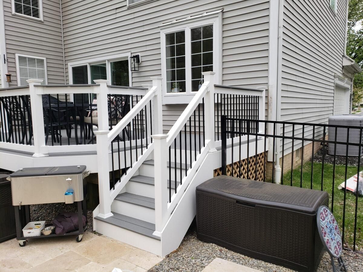 Back deck with white stairs, black railing, wicker bench, and covered grill beside a house