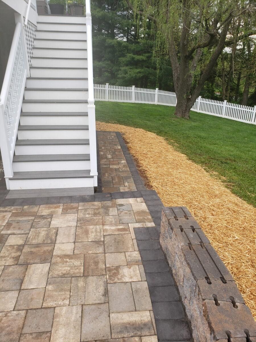 Outdoor staircase beside a paver patio, gravel path, grass, trees, and a white fence.