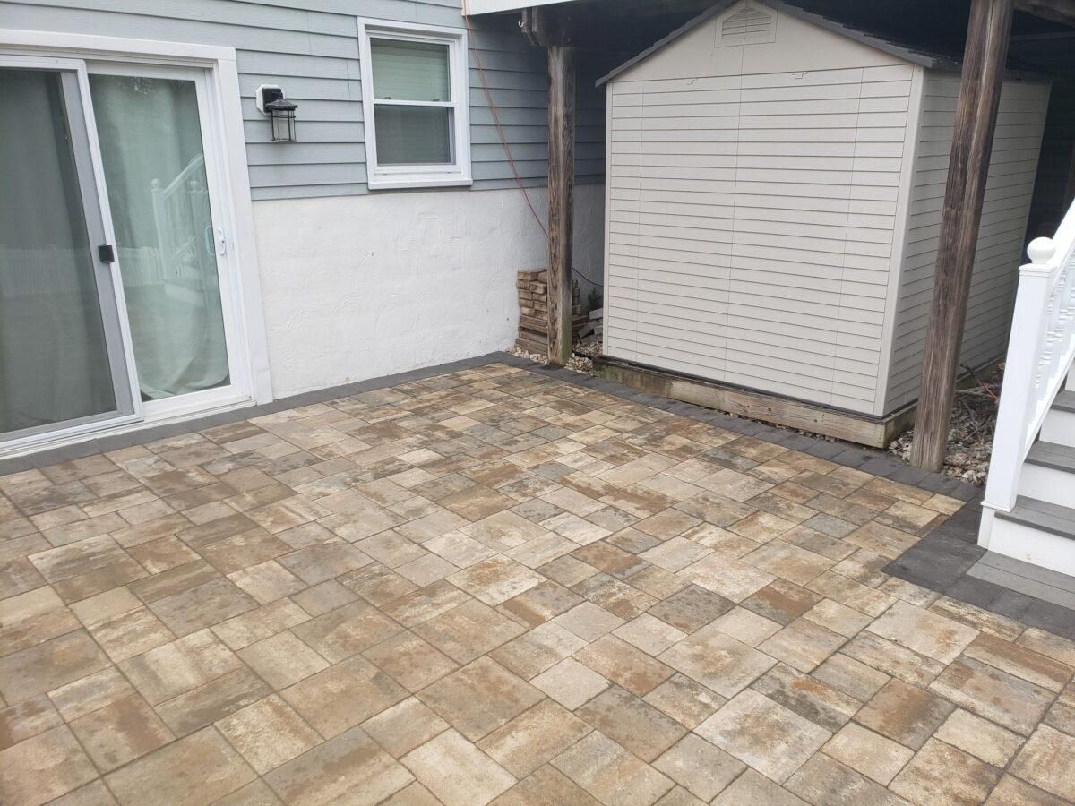 Stone patio beside a house, with sliding glass door, shed, and white stairs.