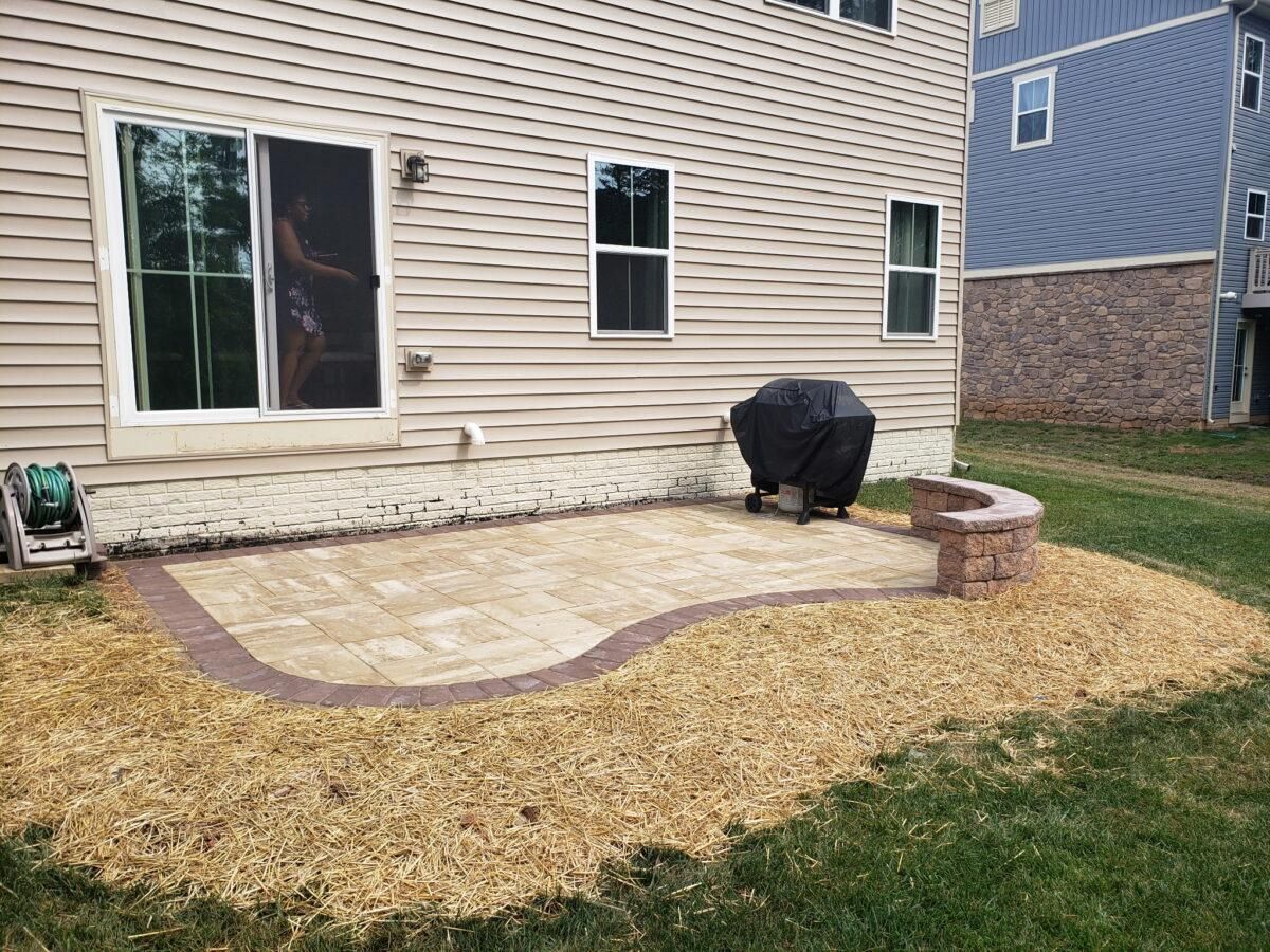 Backyard stone patio beside beige house with sliding glass door, grill, and curved brick fire pit edge
