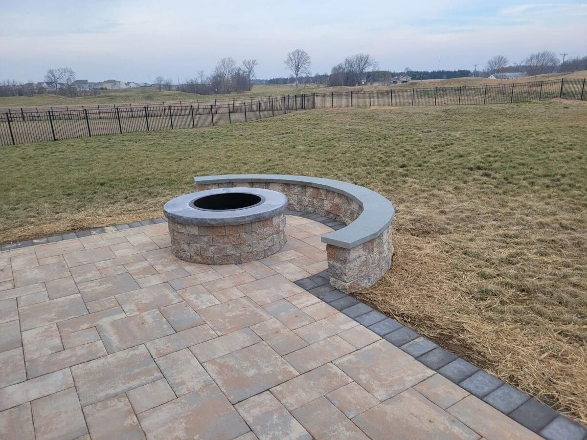 Stone fire pit with curved bench on a brick patio beside an open grassy field
